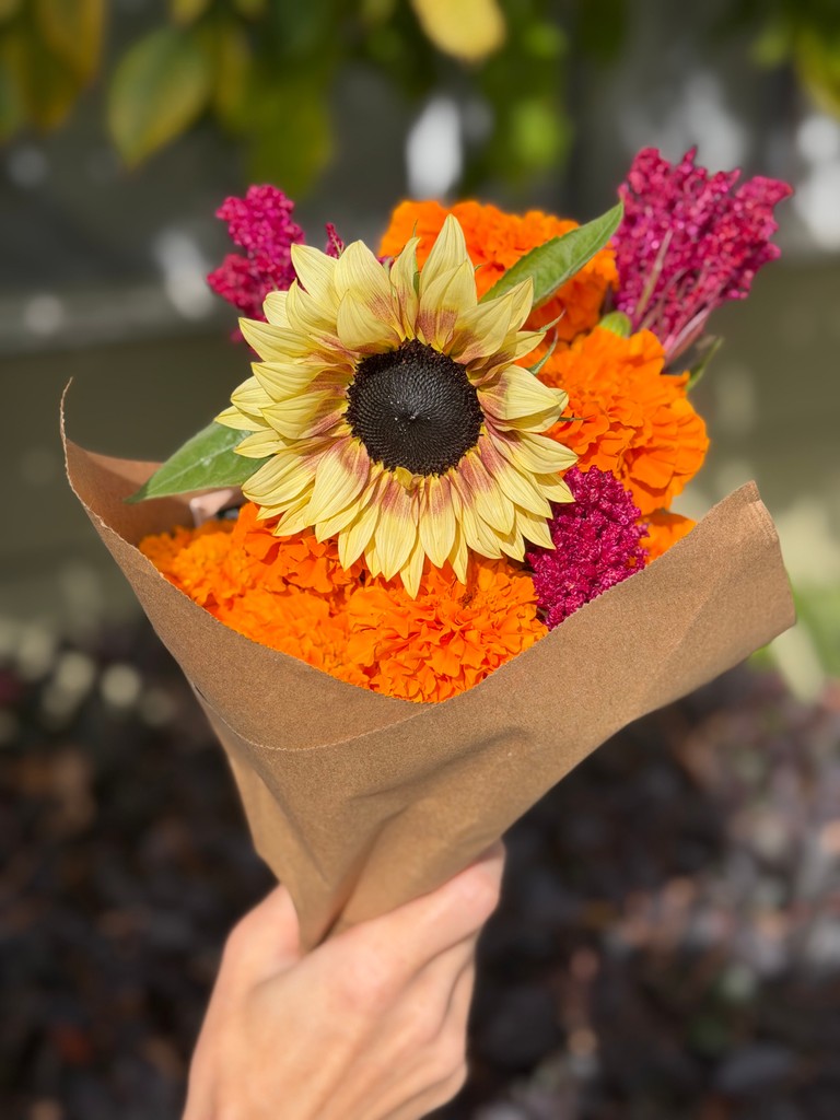 A yellow sunflower with orange and magenta flowers in a brown paper-wrapped bouquet